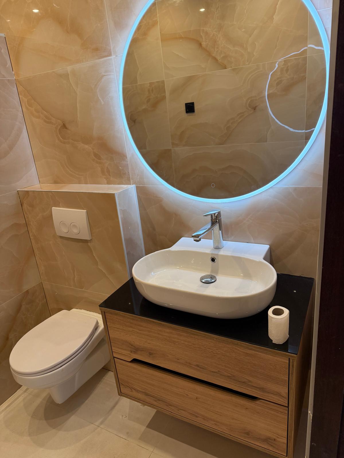Badkamer met ronde ledspiegel, houten wastafelmeubel en zwart werkblad